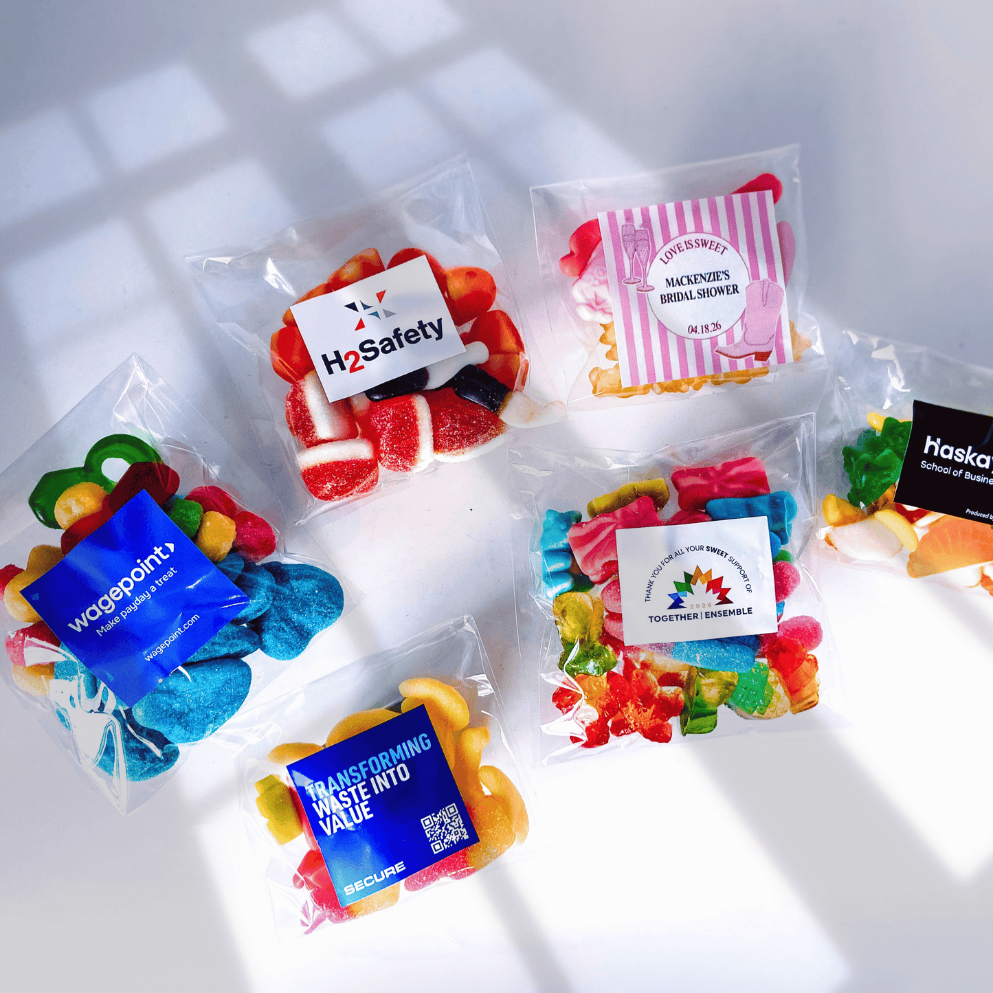 An assortment of clear candy bags filled with various colorful treat mixes, each featuring a custom corporate client logo sticker.