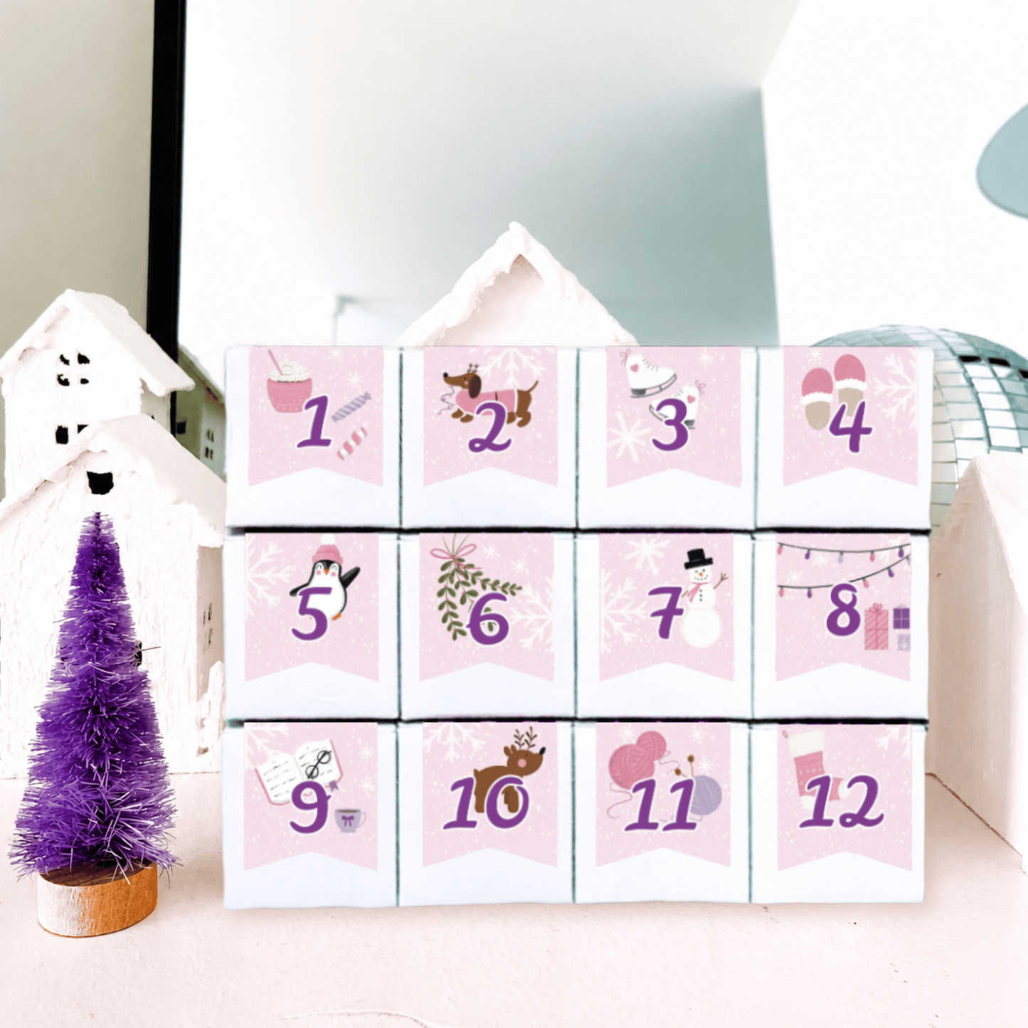 The Sugar Cube 12-Day Family Candy Advent Calendar featuring 12 numbered white candy boxes with pink holiday illustrations, displayed beside a miniature purple Christmas tree and winter decorations.